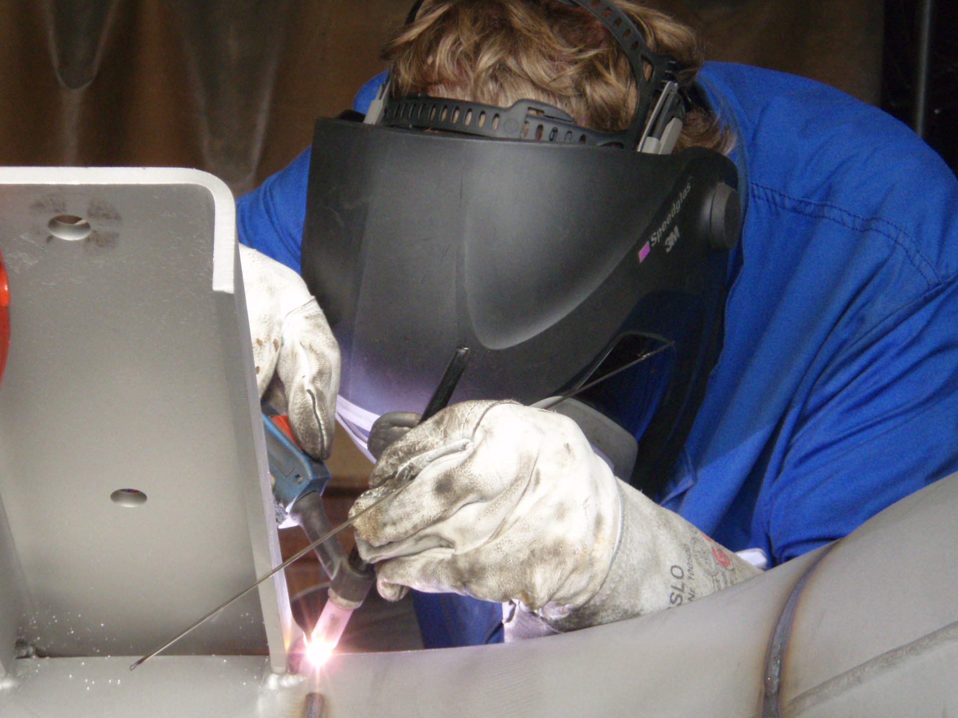 Facharbeiter mit Schweißermaske beim manuellen Schweißen von Metallbauteilen – präzise Handarbeit mit Schutzausrüstung.